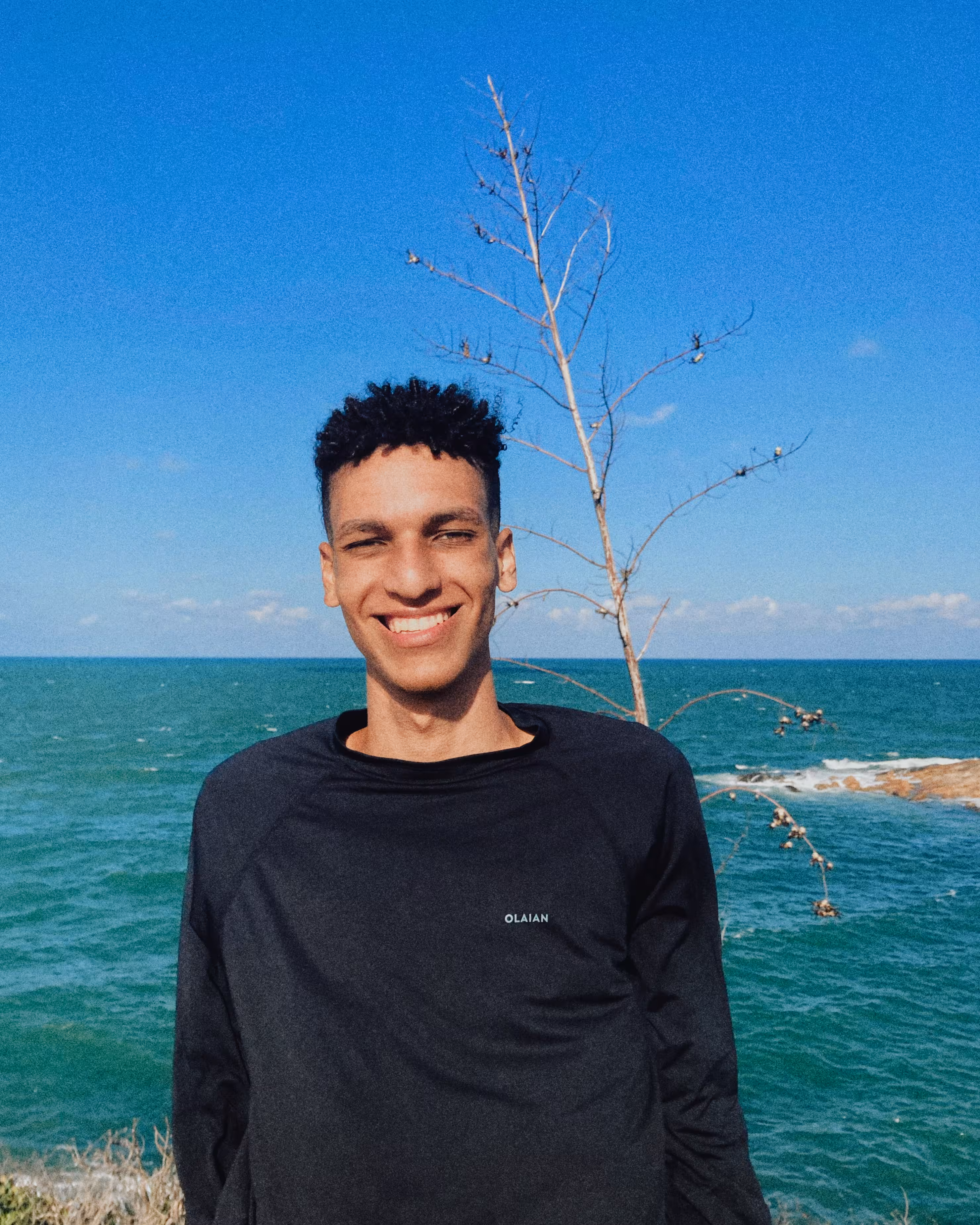 O autor da página vestindo uma camisa UV preta sorri. O mar está ao fundo da imagem.